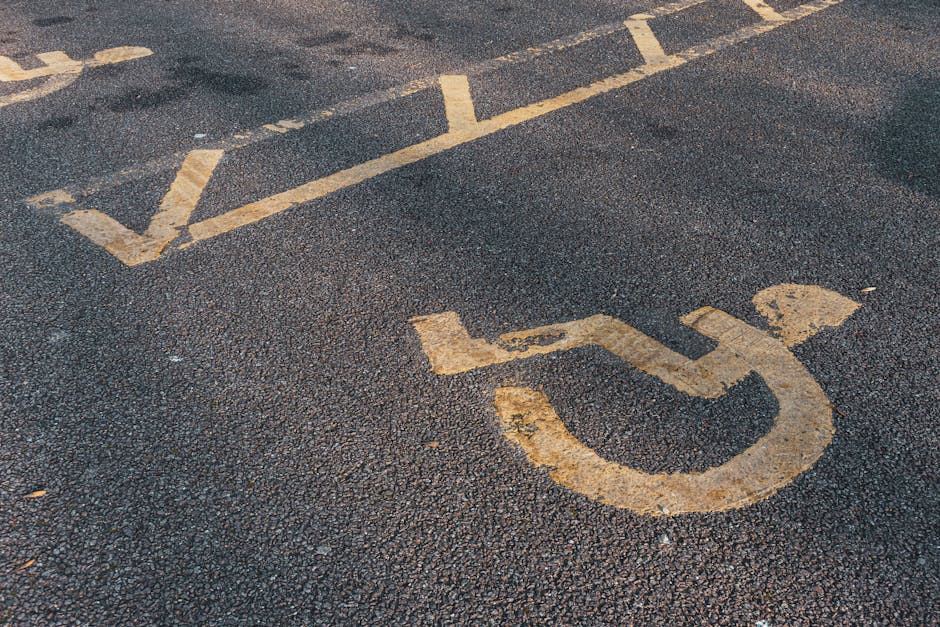 A close-up view of a section of asphalt parking lot pavement featuring a faded yellow wheelchair symbol painted on the surface, indicating designated accessible parking. The textured asphalt has a dark grey, slightly rough finish with small pebbles visibly embedded. The wheelchair icon, positioned towards the lower right, is worn with parts of the paint chipped or faded, revealing the dark asphalt beneath. Above the symbol, several parallel yellow lines run across the pavement, marking the boundary of parking bays. The lines are also weathered, with some areas showing signs of peeling. The overall scene appears to be outdoors under natural light, with no other objects or vehicles visible in the frame. This image could relate to accessible parking spaces managed through private arrangements or independent clearance services such as those offered by Rubbish Clearance Wandsworth, emphasizing the importance of clear and compliant parking markings for waste collection or service access.