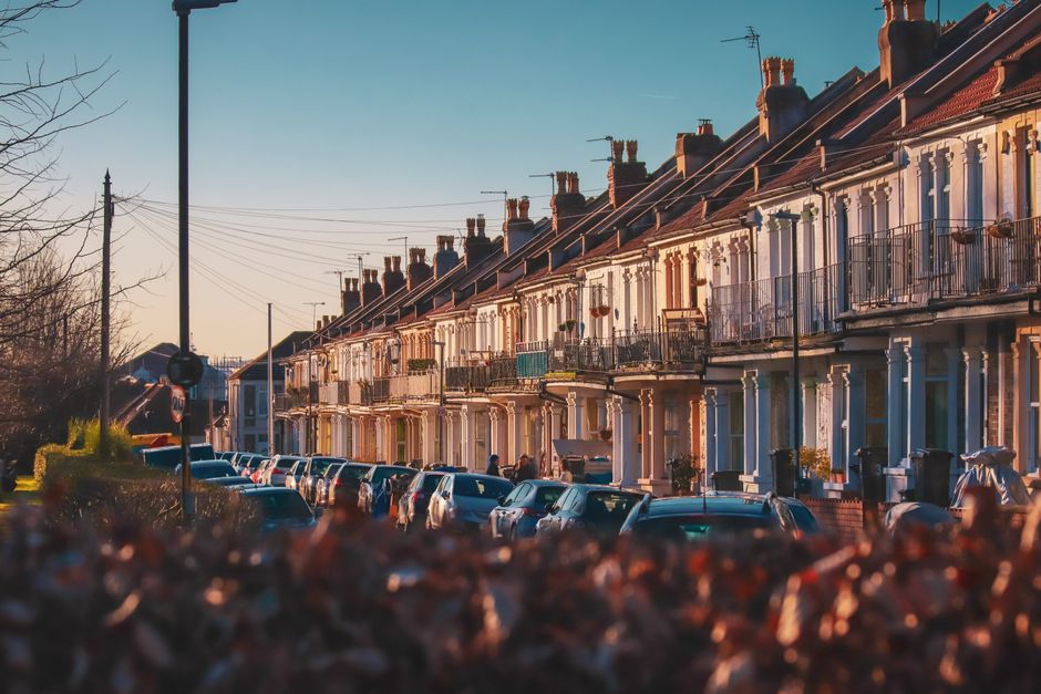 The image depicts a residential street lined with a row of terraced houses featuring brick and plaster facades in light pastel shades, mainly white and cream, with some retaining a slightly weathered appearance. Each house has a small front balcony with black wrought iron railings, and several display potted plants or outdoor furniture along the front. The street is filled with parked cars, including various hatchbacks, sedans, and small SUVs, arranged in parallel along the curb on both sides, with some vehicles partially obscured by a foreground pile of blurred rubbish or debris. A tall lamppost stands on the left side of the image, and the background shows a clear sky with warm natural lighting indicating either early morning or late afternoon, casting gentle shadows and a slightly golden hue over the scene. The environment suggests a typical urban or suburban neighbourhood, with no visible signs of active rubbish collection or disposal activities, aligning with independent collection or private waste handling scenarios often associated with residential property management or rubbish removal services like Rubbish Clearance Wandsworth, who may handle such waste accumulation in similar settings.