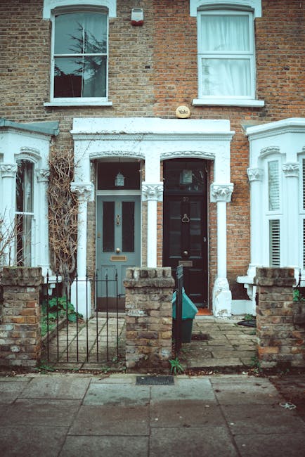 The image depicts the front facade of two adjacent terraced houses with brick exteriors, typical of residential properties in Wandsworth. The left house features a grey painted door with a vertical frosted glass panel, set within a white decorative porch with intricate plasterwork and two small columns supporting a curved pediment. Above, there is a single-pane sash window with a white frame, showing a reflection of a tree in the glass. The right house has a black door with a brass door knocker and letterbox, also within a white decorative porch with similar plaster details, and above it, a sash window with white frames and closed curtains. Both properties are separated by a narrow paved pathway, bordered by low brick walls with metal railings on top, and small gates leading to the front entrances. The front gardens are minimal, with some sparse greenery along the base of the walls. An outdoor light fixture is visible on the porch of the left house. This scene illustrates typical urban residential architecture and the potential for private waste collection or rubbish clearance services in such properties, with a focus on the immediate external environment of these terraced homes.