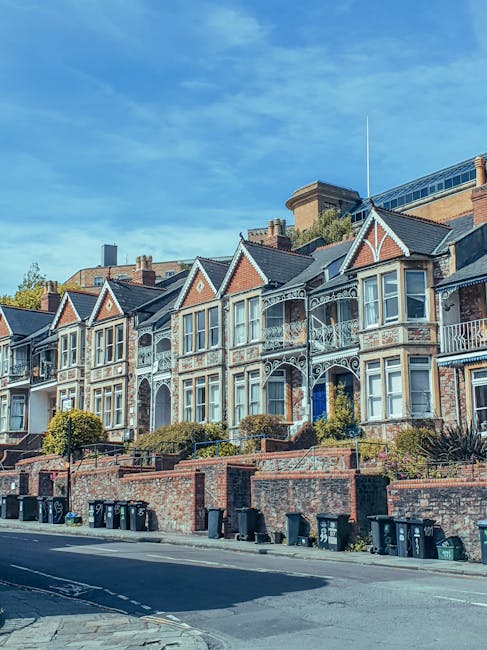 The image depicts a residential street lined with a row of terraced houses featuring brick and plaster facades in light pastel shades, mainly white and cream, with some retaining a slightly weathered appearance. Each house has a small front balcony with black wrought iron railings, and several display potted plants or outdoor furniture along the front. The street is filled with parked cars, including various hatchbacks, sedans, and small SUVs, arranged in parallel along the curb on both sides, with some vehicles partially obscured by a foreground pile of blurred rubbish or debris. A tall lamppost stands on the left side of the image, and the background shows a clear sky with warm natural lighting indicating either early morning or late afternoon, casting gentle shadows and a slightly golden hue over the scene. The environment suggests a typical urban or suburban neighbourhood, with no visible signs of active rubbish collection or disposal activities, aligning with independent collection or private waste handling scenarios often associated with residential property management or rubbish removal services like Rubbish Clearance Wandsworth, who may handle such waste accumulation in similar settings.