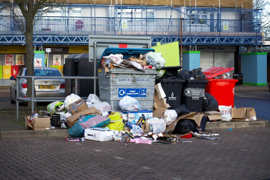 A large collection of mixed waste and rubbish is piled on a paved parking area in front of a waste collection point, including black and red plastic garbage bags, flattened cardboard boxes, crumpled paper, and various packaging materials. The rubbish spills over the top of a grey recycling bin labeled for mixed paper and cardboard, with its lid partially open. Adjacent to the recycling bin, there are smaller black bins, with one black bin appearing to contain additional waste, and a red bin that might be designated for general waste disposal. To the left, a metallic vehicle with a rear license plate is parked near a metal safety railing that separates the waste area from the paved sidewalk. Behind the waste scene, a row of shops with large windows and signage is visible, partially obscured by a large tree trunk on the left. In the background, a multi-storey building with scaffolding and safety netting indicates ongoing construction or renovation work. The daylight suggests a cloudy day, creating even, diffuse lighting across the scene, highlighting the cluttered appearance typical of an informal rubbish collection or overflow situation that might be addressed through private waste removal services, as provided by Rubbish Clearance Wandsworth.