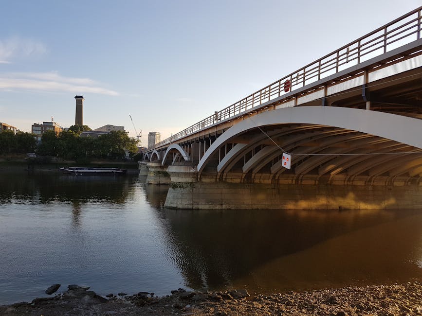 A wide-angle view of a large white arch bridge spanning across a calm body of water during daylight hours, showing the underside with visible support structures and the upper deck equipped with red railings. In the background, there are several modern buildings, including one with a tall chimney and a construction crane, suggesting ongoing development. The water reflects the bridge and surrounding scenery, with some ripples near the shoreline composed of small rocks and gravel. The scene is illuminated by natural light, with a partly cloudy sky overhead. This environment is characteristic of urban riverside areas where private or independent waste removal services, such as those provided by Rubbish Clearance Wandsworth, may operate in close proximity to infrastructure and residential zones, supporting effective rubbish disposal and clearance activities in the vicinity.
