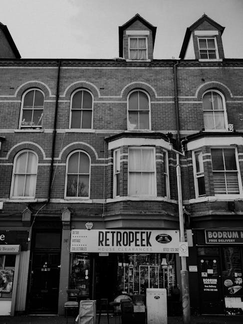 A black and white photograph of a multi-storey brick building with three floors, featuring arched and rectangular windows with white frames. The ground floor contains a retail business named 'Retro PeeK,' which offers house clearances, indicated by a large sign above the entrance. The storefront has glass doors and windows displaying various items, possibly related to waste or clearance services. To the right, part of another shop's signage, 'Bodrum,' can be seen, suggesting commercial activity in the area. In front of the building, on the pavement, there are two small barrels or bins and a large trash or recycling container, hinting at waste collection or clearance activities. The overall scene appears to be in an urban commercial district, possibly prepared for or in the process of rubbish removal or private waste handling. The lighting is natural, with overcast skies casting even illumination across the scene, providing a neutral, observational view of the premises that aligns with waste management and rubbish clearance contexts.